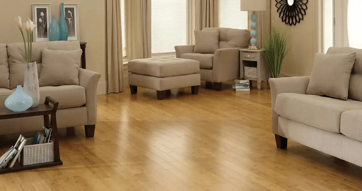 A living room with professionally installed bamboo flooring, beige sofas, a matching ottoman, a side table with a blue lamp, vases, framed pictures, and large windows with blinds and curtains in Manchester, NH